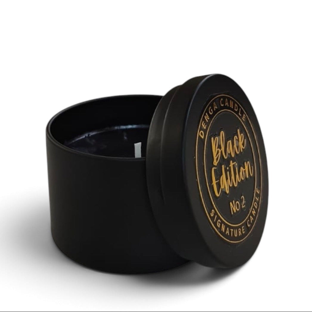 A black candle tin with its lid leaning against the container, showing a small white wick inside. The lid is printed with gold lettering that reads 'DENGA CANDLE Black Edition No2