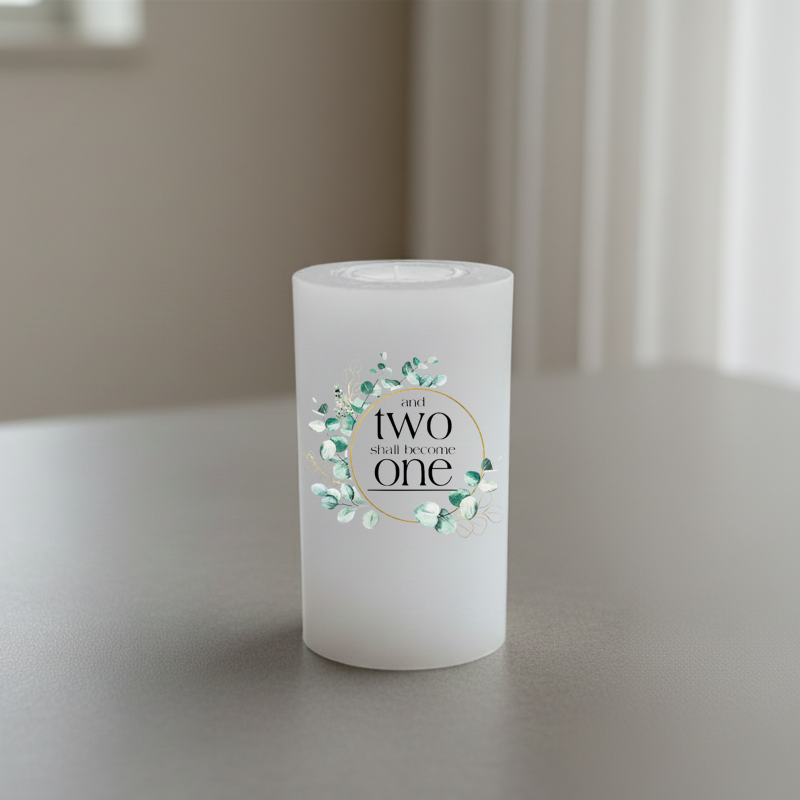 A white candle with text surrounded by a circular floral wreath in Eucalyptus tones, set on a grey tabletop with a blurred indoor background.