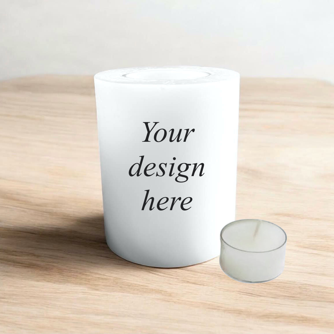 A white candle with the text 'Your design here' printed in black, set on a light wood surface against a neutral background.