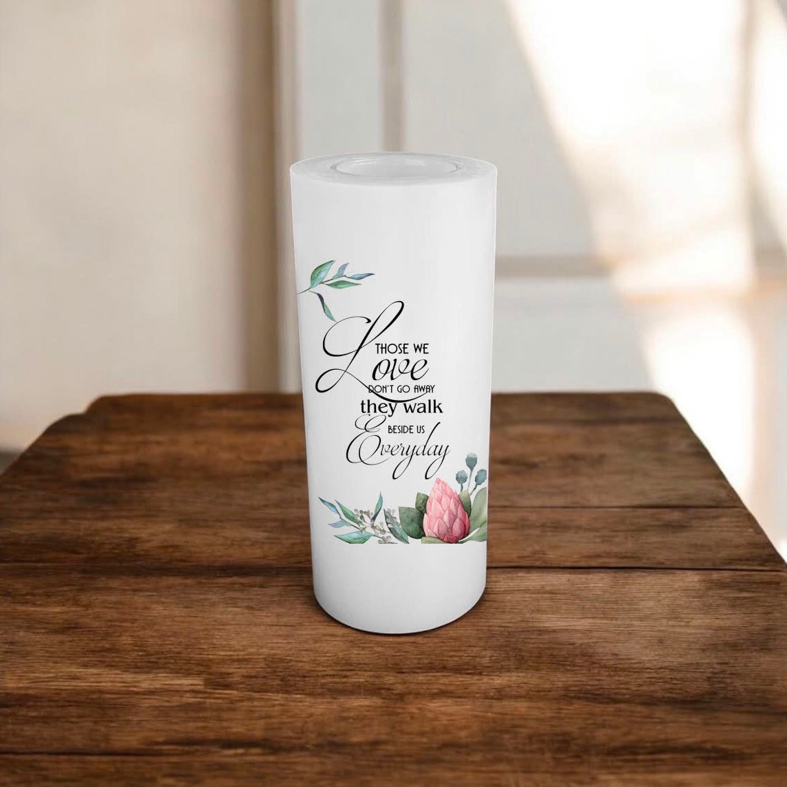 A white candle with a printed quote and floral illustration, standing on a rustic wooden surface against a softly blurred indoor background.