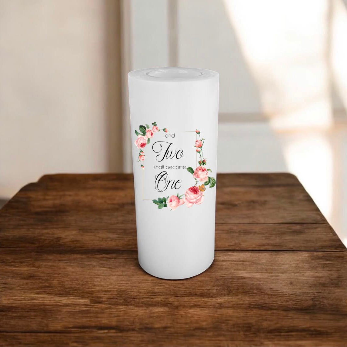 A white candle with a floral and script design reading 'and Two shall become One,' displayed on a wooden surface in a softly lit indoor setting.