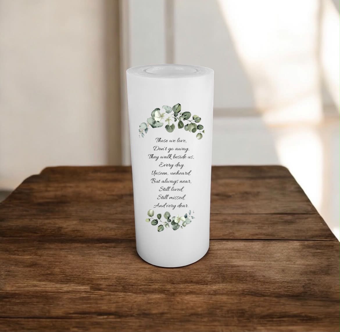 A white candle with printed black script text and green floral illustrations, standing on a wooden surface against a softly blurred indoor background.