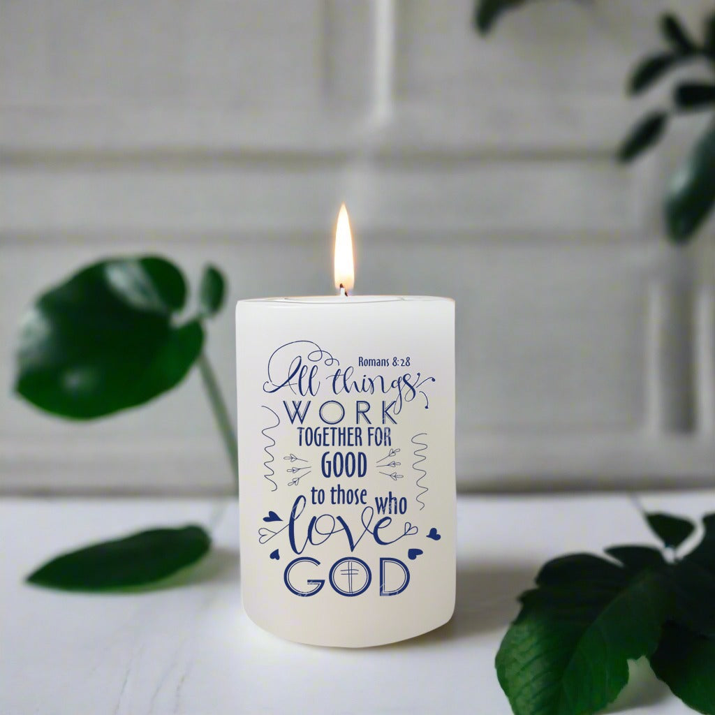 Candle with text 'All things work together for good to those who love God' on a white surface with green leaves.