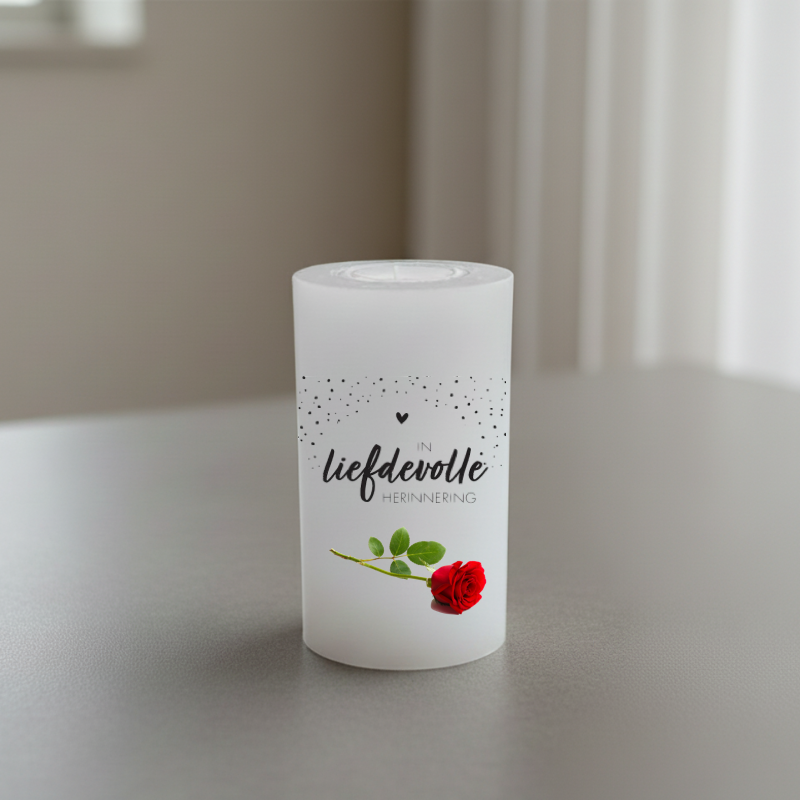 A white candle with black Afrikaans text reading and a red rose illustration, on a grey surface