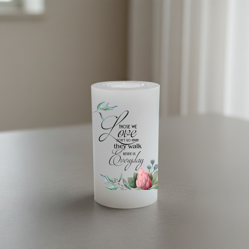 A white candle printed black script text and pink floral illustrations, standing on a grey tabletop.