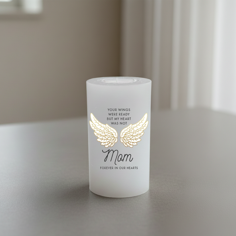 A white memorial candle with a gold angel wings graphic and the word 'Mom' in black script.