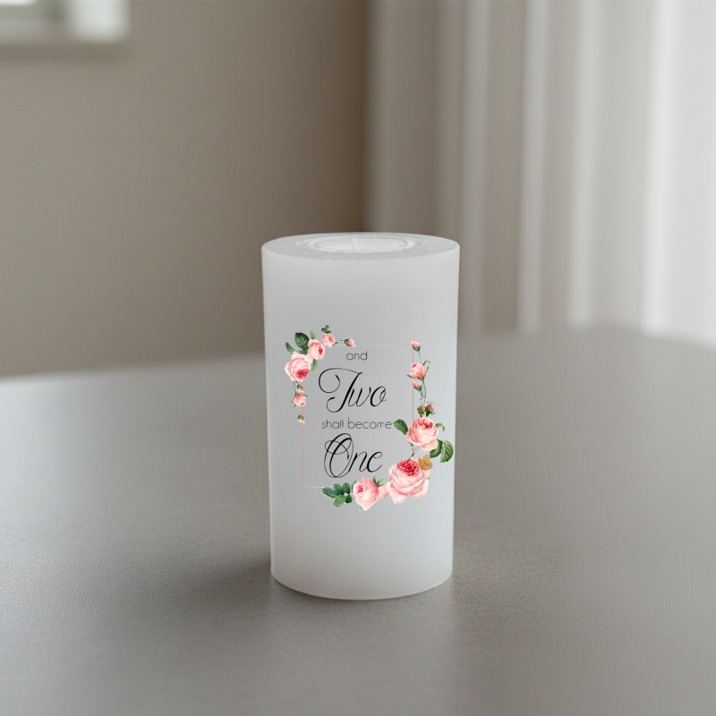 A white cylindrical candle with floral design and the words 'and two shall become One' in black script, shown on a tabletop in a softly lit indoor setting.