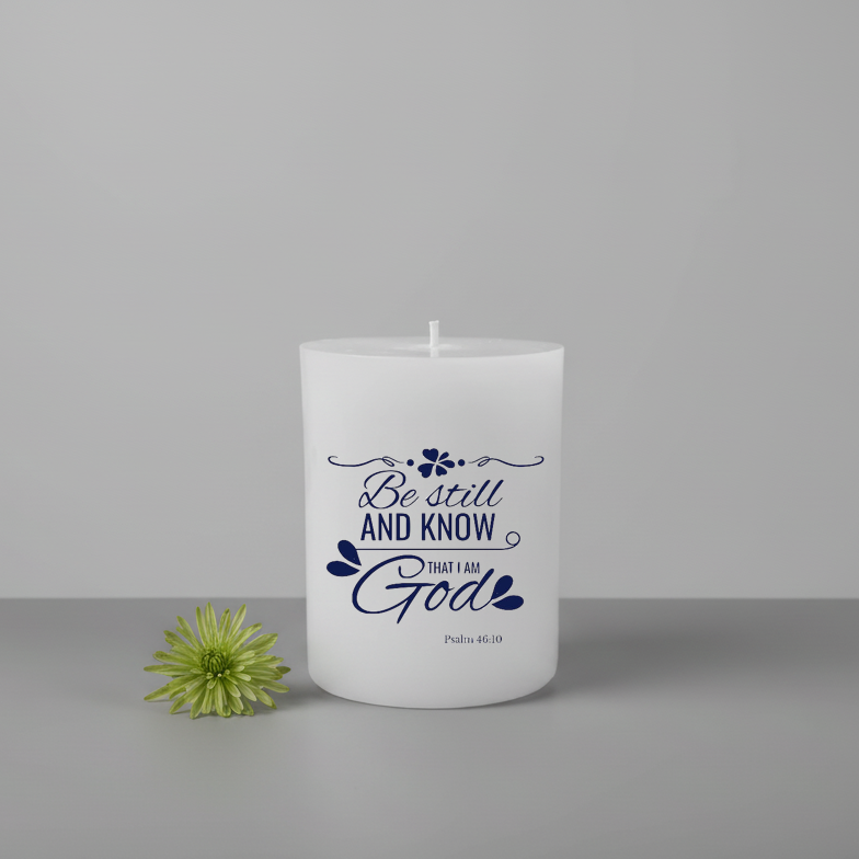 White candle with blue text 'Be still and know that I am God' next to a green flower on a gray background