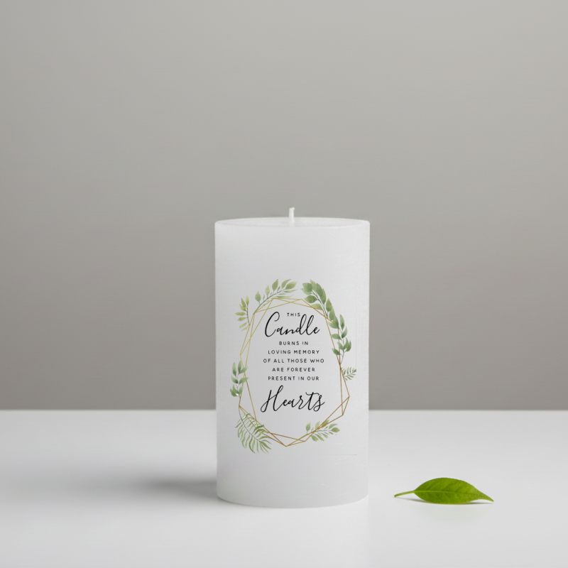 A white memorial candle with a printed green floral wreath and gold geometric border, and a green leaf beside it.