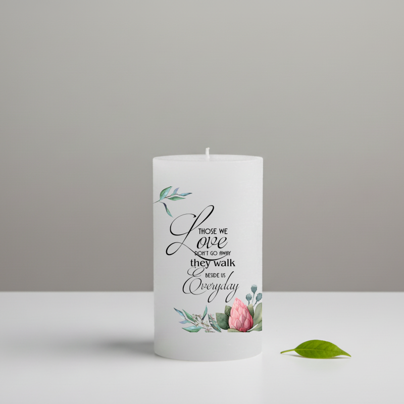 A white candle printed black script text and pink floral illustrations, standing on a white surface and green leaf beside