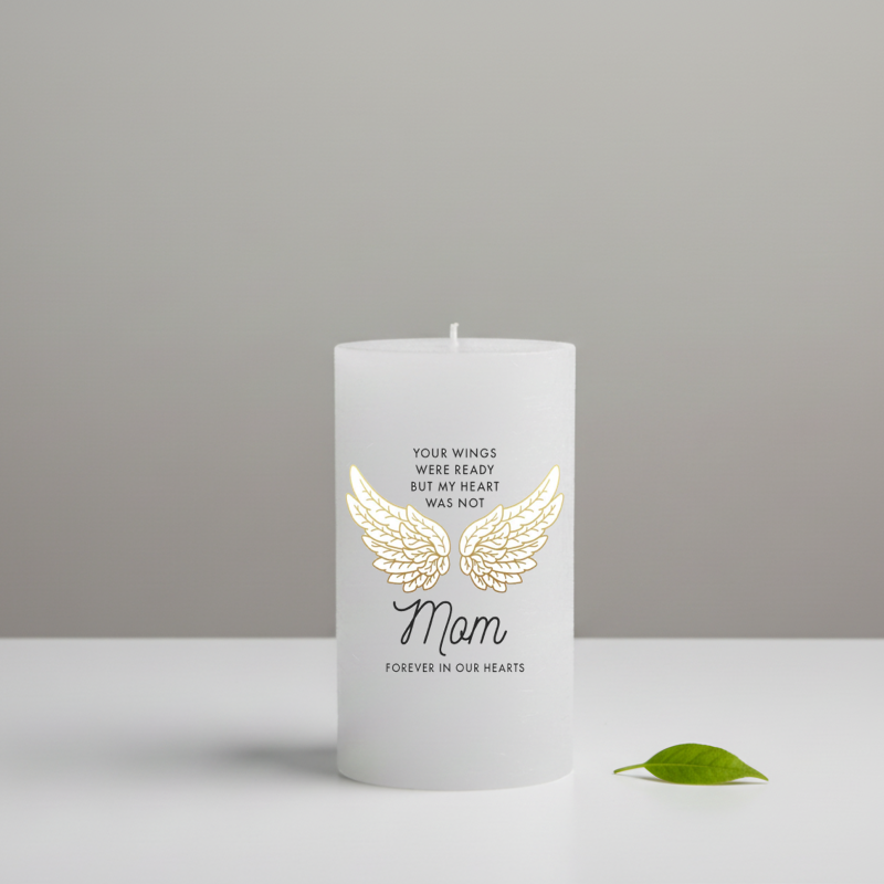 A white candle with a printed gold angel wings design and the word 'Mom', shown on a light grey background with a small green leaf beside it.