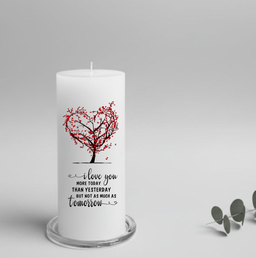 A white candle featuring a red heart-shaped tree design and the printed message 'I love you MORE TODAY THAN YESTERDAY BUT NOT AS MUCH AS tomorrow'.