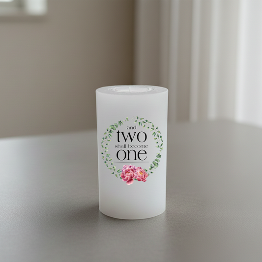 A white candle with a printed graphic that reads 'and two shall become one' in black script, surrounded by a green floral wreath and pink roses.