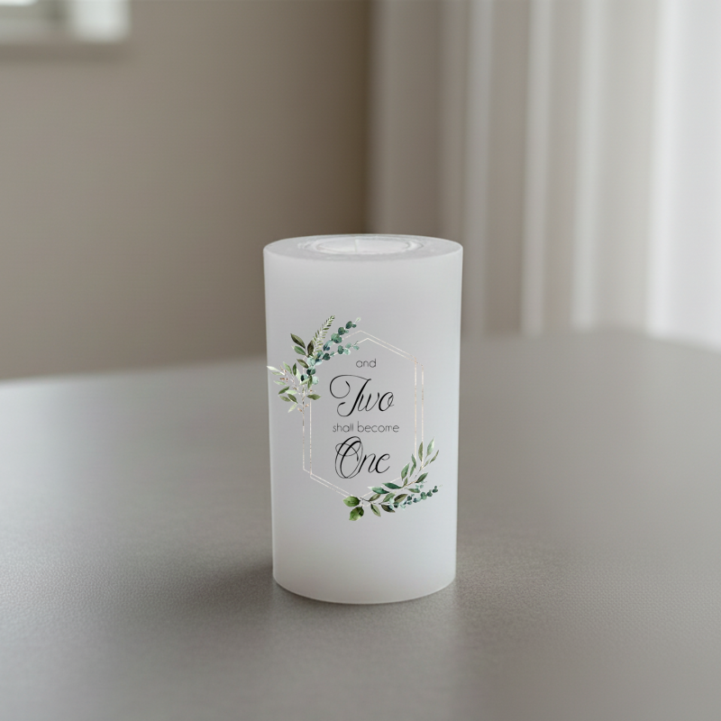 A white candle with a botanical floral design and the printed words 'and Two shall become One' in black script, set on a neutral grey surface with a soft-focus background.