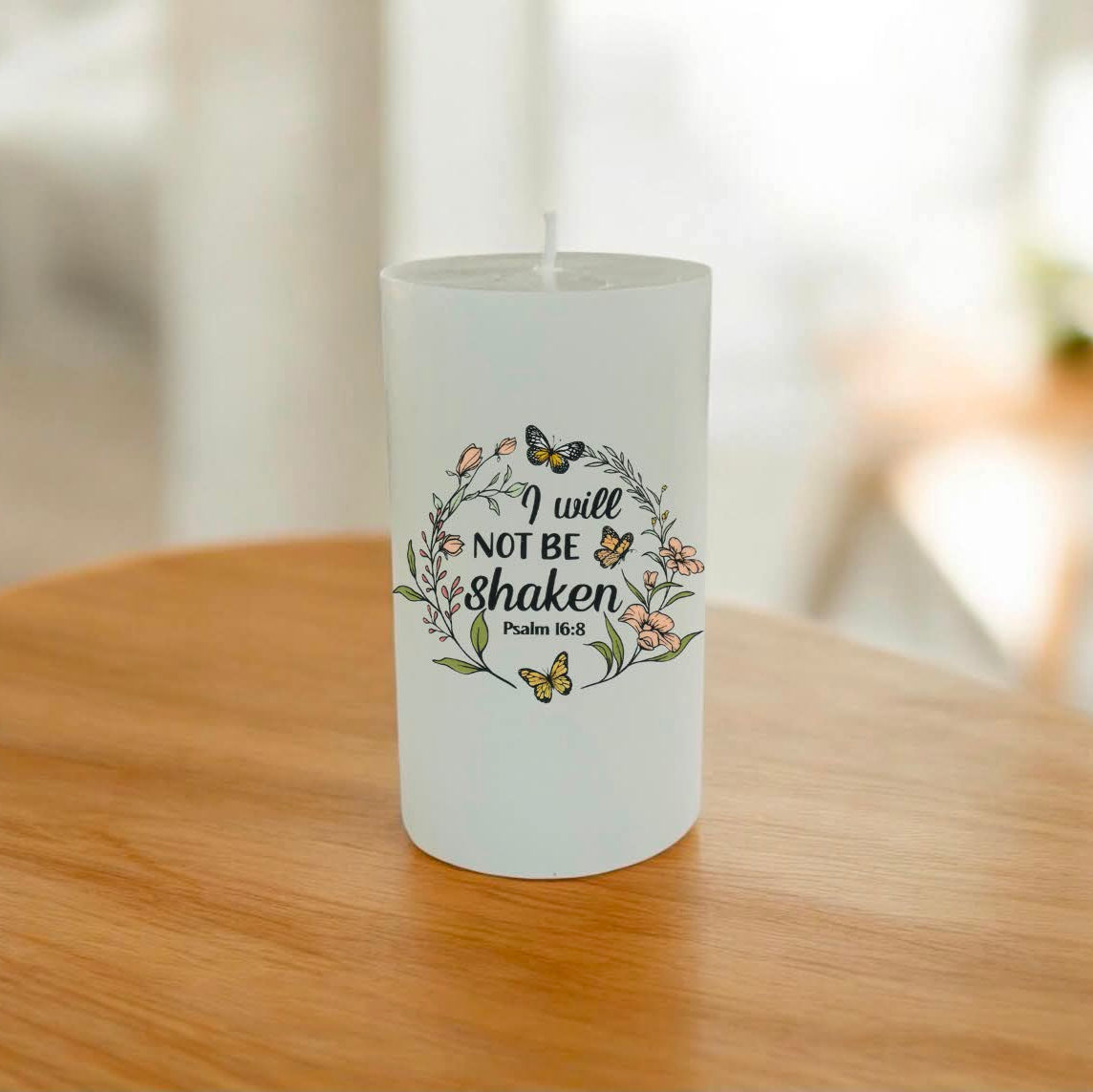 A white candle with a printed Bible verse and floral illustrations, sitting on a wooden table with a blurred indoor background.