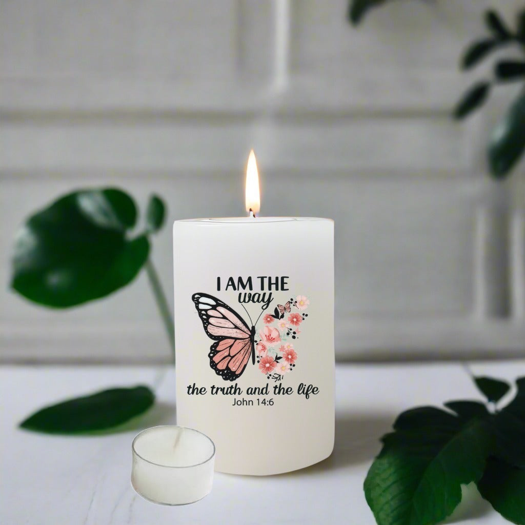 A white pillar candle with a lit flame, featuring a printed butterfly and pink floral design with the text 'I AM THE way the truth and the life John 14:6'.