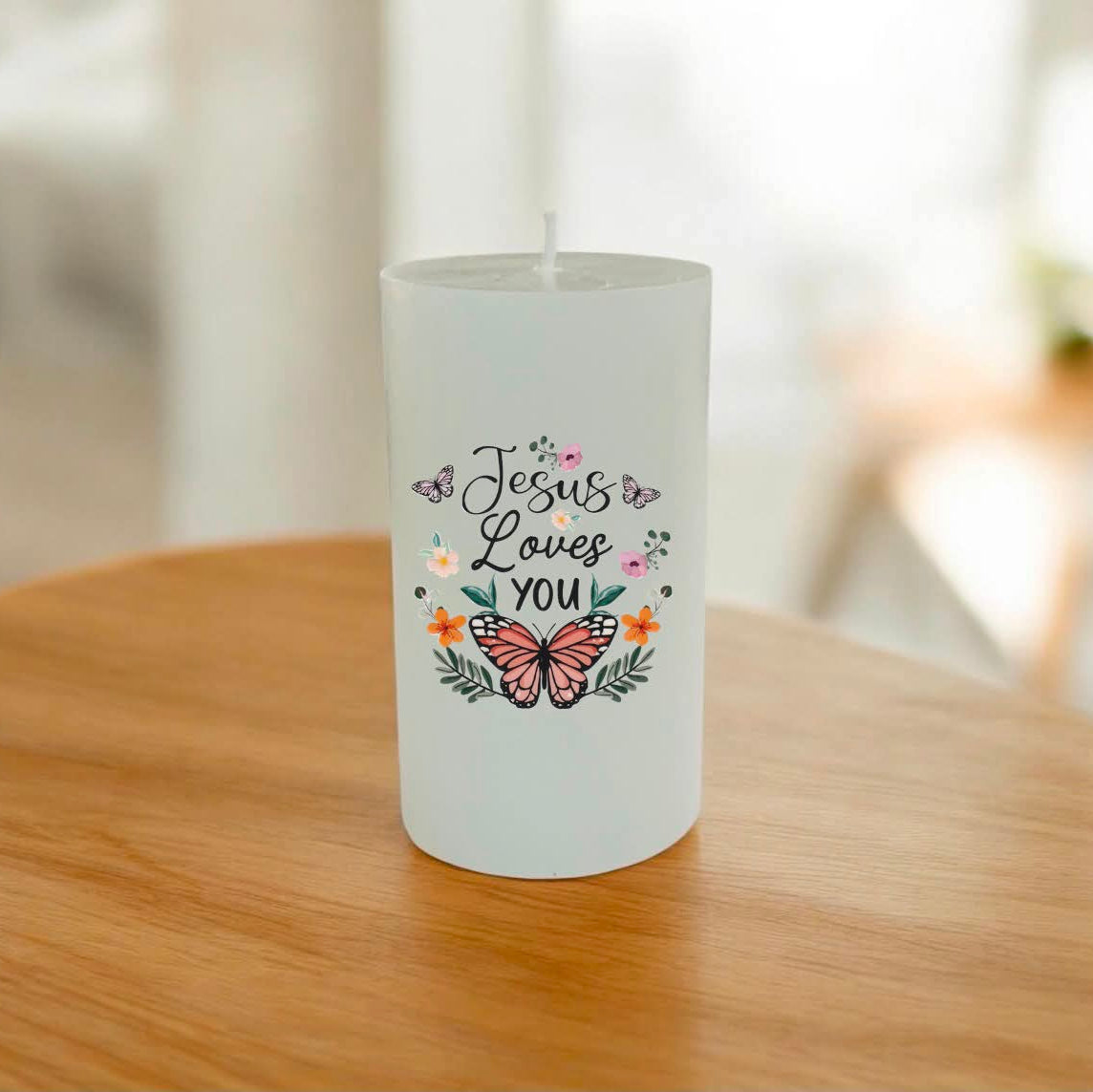 A white candle with a printed design that reads 'Jesus Loves You' in black script, decorated with a pink butterfly, small flowers, and green leaves.