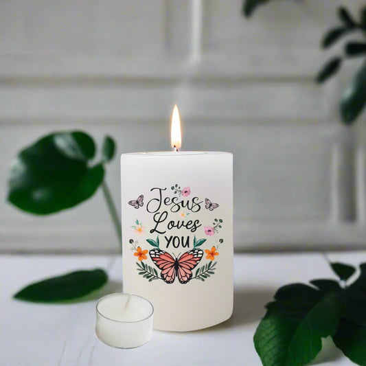 A white pillar candle with a lit flame, featuring the text 'Jesus Loves you' in black script with a pink butterfly and small floral illustrations. 
