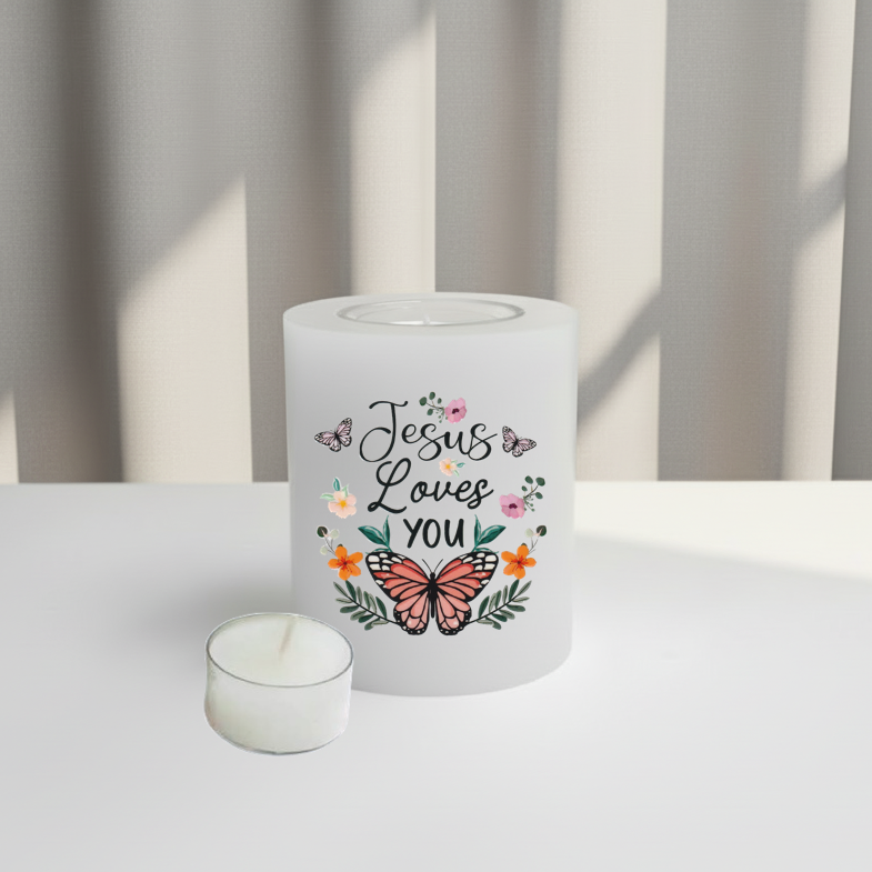 A white candle holder with the words 'Jesus Loves YOU' and a butterfly design, displayed on a white surface with soft window light and curtains in the background.