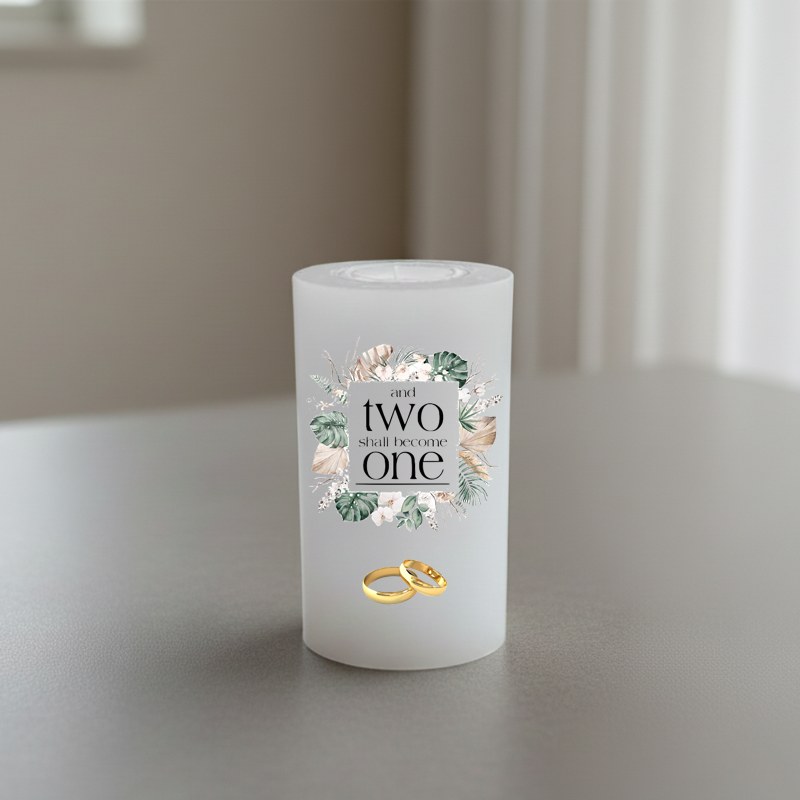 white candle with a floral graphic and the text 'and two shall become one' on the front, shown standing on a grey surface with a softly blurred indoor background.
