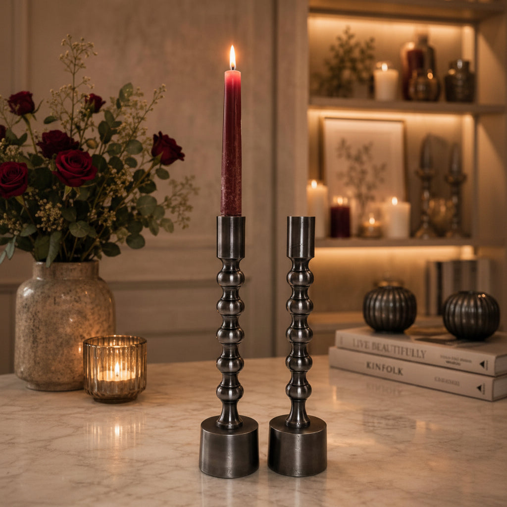 Two dark metal candlestick holders on a marble table, one holding a lit Ruby taper candle and the other empty, with a warm, cozy interior background