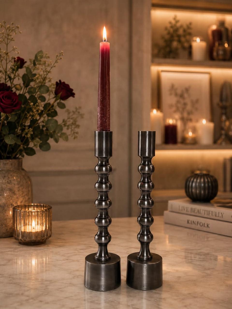 Two dark metal candlestick holders on a marble table, one holding a lit dark red taper candle and the other empty, with a warm, cozy interior background