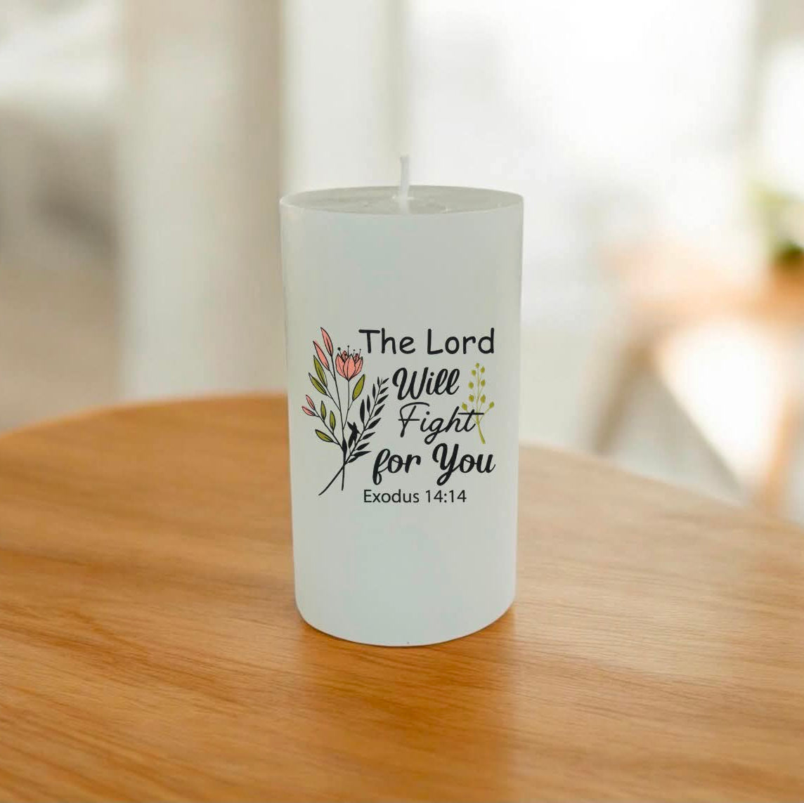A white pillar candle with a printed religious quote and floral illustration, sitting on a wooden table in a softly blurred indoor setting.