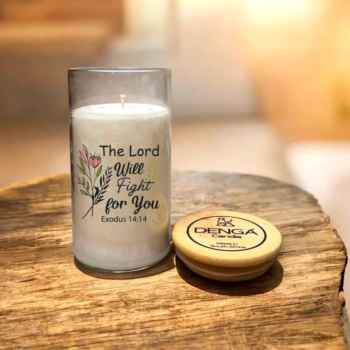 A clear glass jar candle with a printed religious quote and floral illustration sits on a rustic wooden surface next to its round wooden lid.
