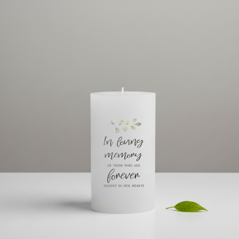 A white pillar candle printed with memorial text, placed on a white surface and a neutral background. 