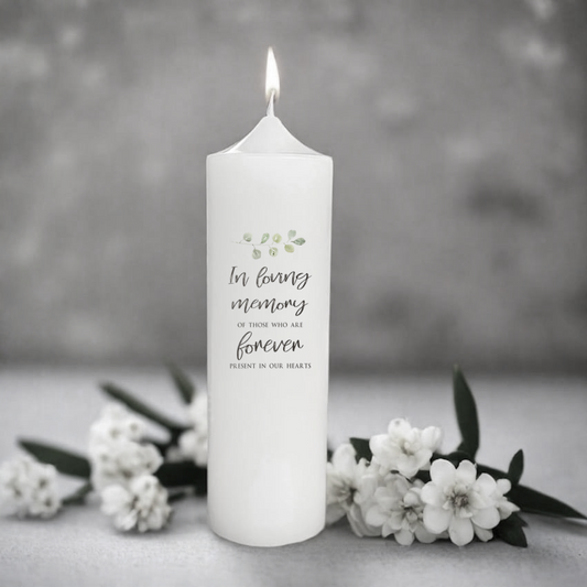 White memorial candle with text 'In loving memory of those who are forever present in our hearts' on a gray background with flowers.
