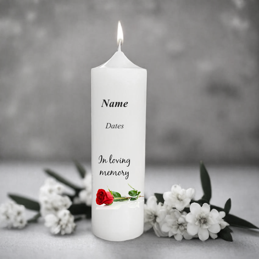 White memorial candle with text and a red rose design, surrounded by white flowers on a gray background