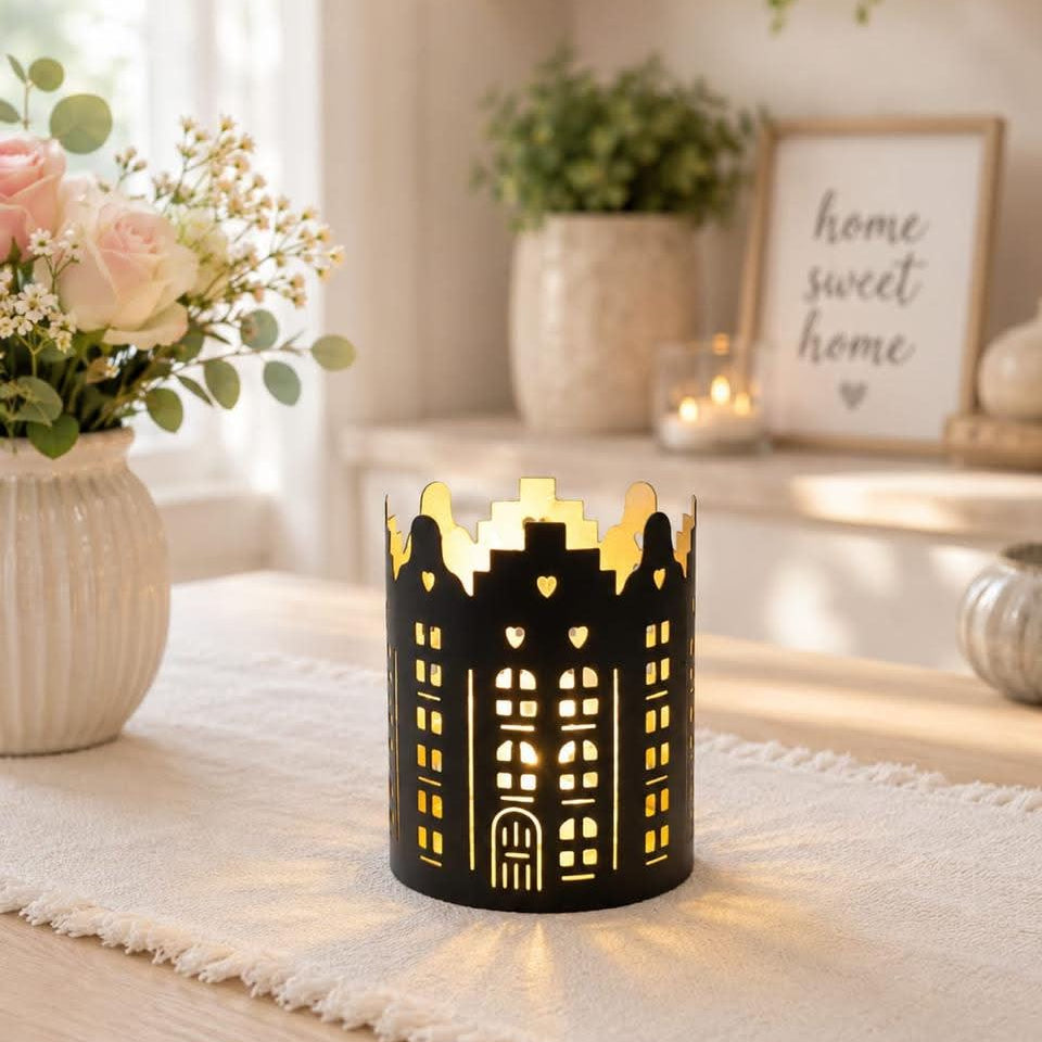A black metal candle holder shaped like a building with heart-shaped windows, lit from within to cast a warm glow, set on a white runner in a cozy, sunlit room.