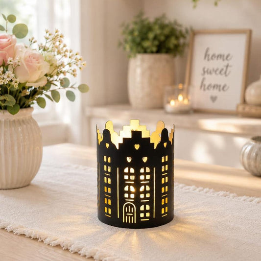 A black metal candle holder shaped like a building with heart-shaped windows, lit from within to cast a warm glow, set on a white runner in a cozy, sunlit room.