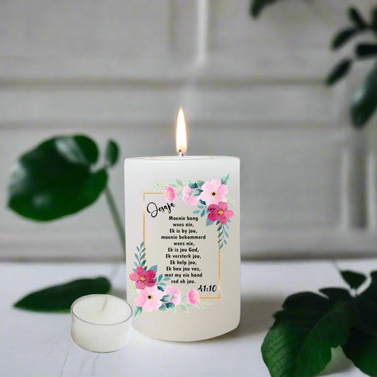 Candle with floral design and Afrikaans text on a white surface with green leaves.