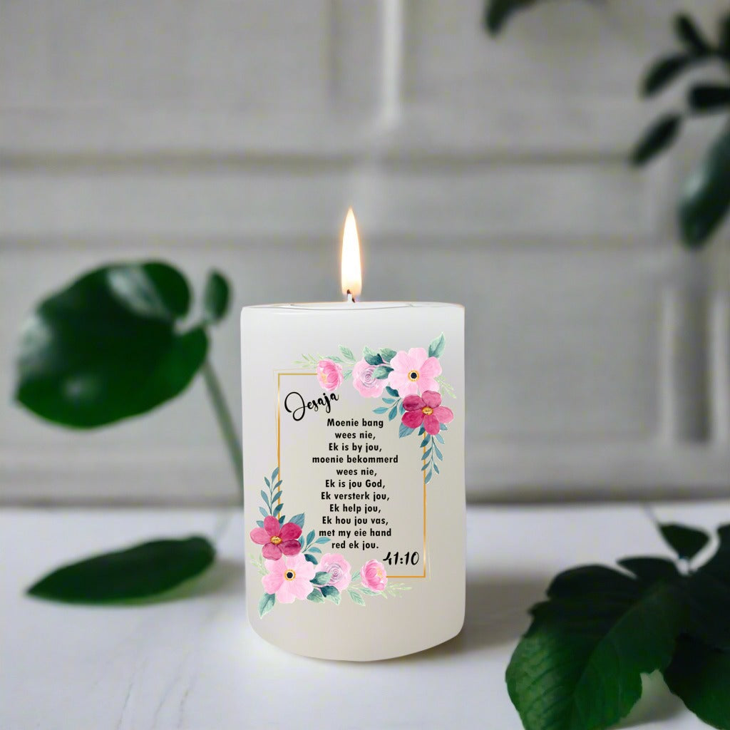 Candle with floral design and Afrikaans text on a white surface with green leaves.