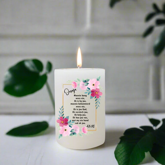 Candle with floral design and Afrikaans text on a white surface with green leaves.