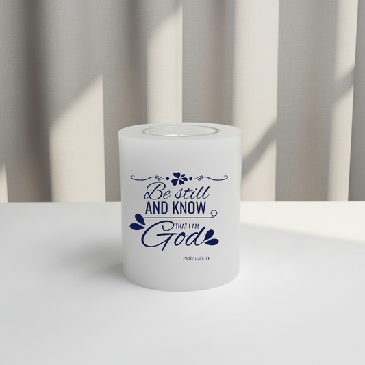 White candle with blue text 'Be still and know that I am God' on a white surface with a neutral background