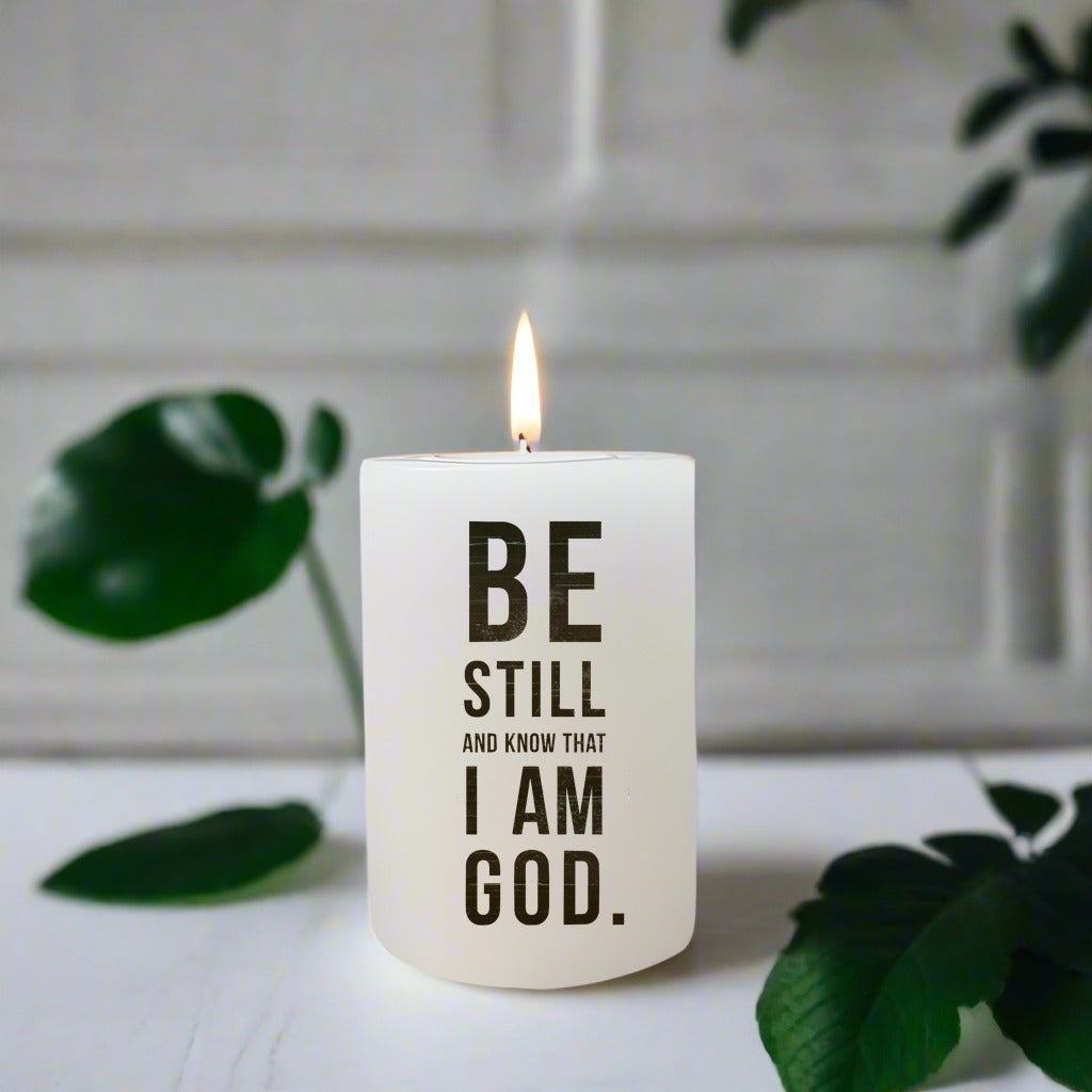 Candle with 'Be Still and Know That I Am God' text on a white surface with green leaves.