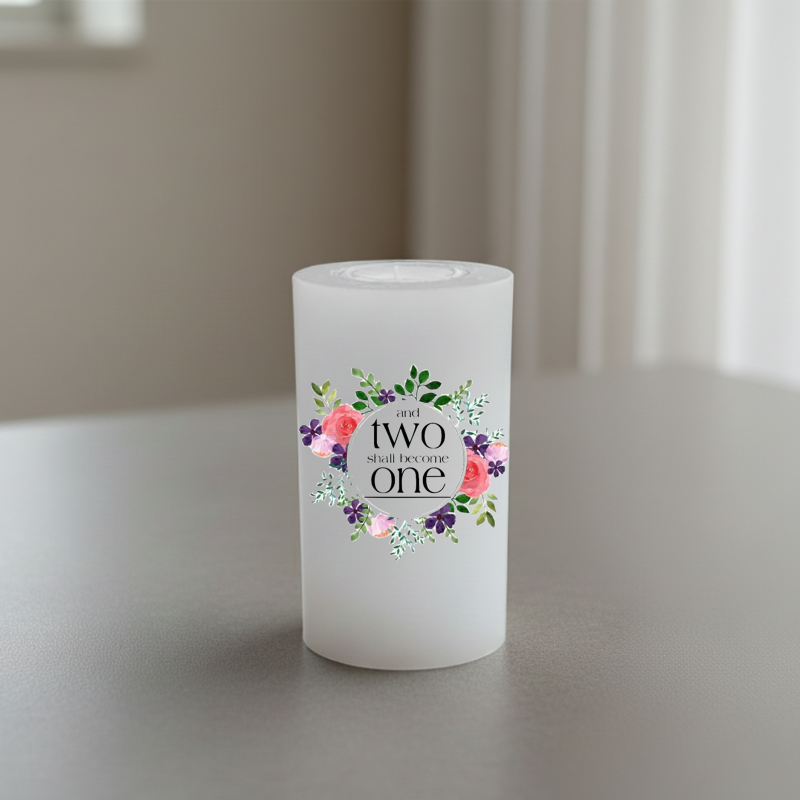 A white cylindrical candle standing on a tabletop, printed in black script and surrounded by a floral wreath in pink, purple, green, and white.