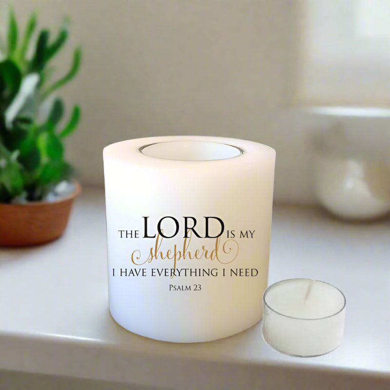A white candle with the printed text 'THE LORD IS MY Shepherd I HAVE EVERYTHING I NEED Psalm 23' in black and gold lettering, shown on a white surface next to a small tea-light candle. 