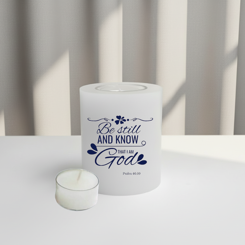 White candle with blue text 'Be still and know that I am God' on a white surface with a neutral background