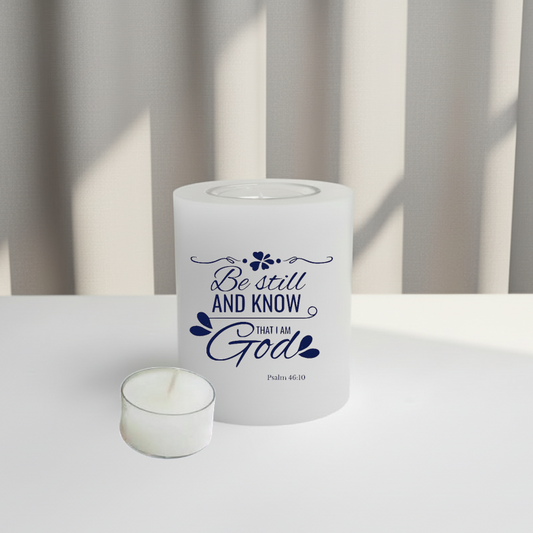 White candle with blue text 'Be still and know that I am God' on a white surface with a neutral background