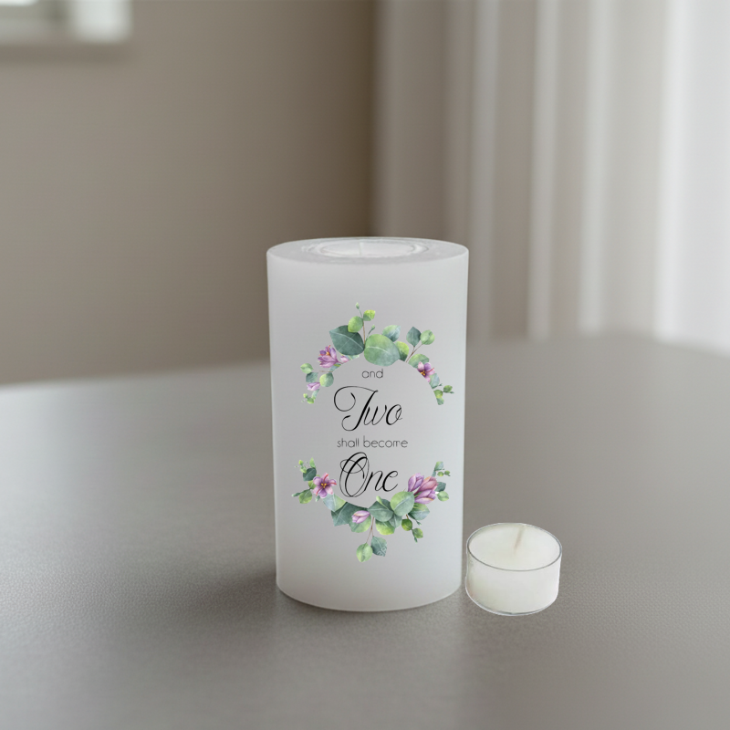 A white candle with a floral design and the words 'and Two shall become One' in black script.