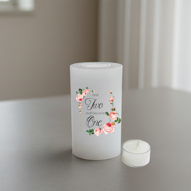 A white candle with floral design and the words 'and two shall become One' in black script, shown on a tabletop in a softly lit indoor setting.