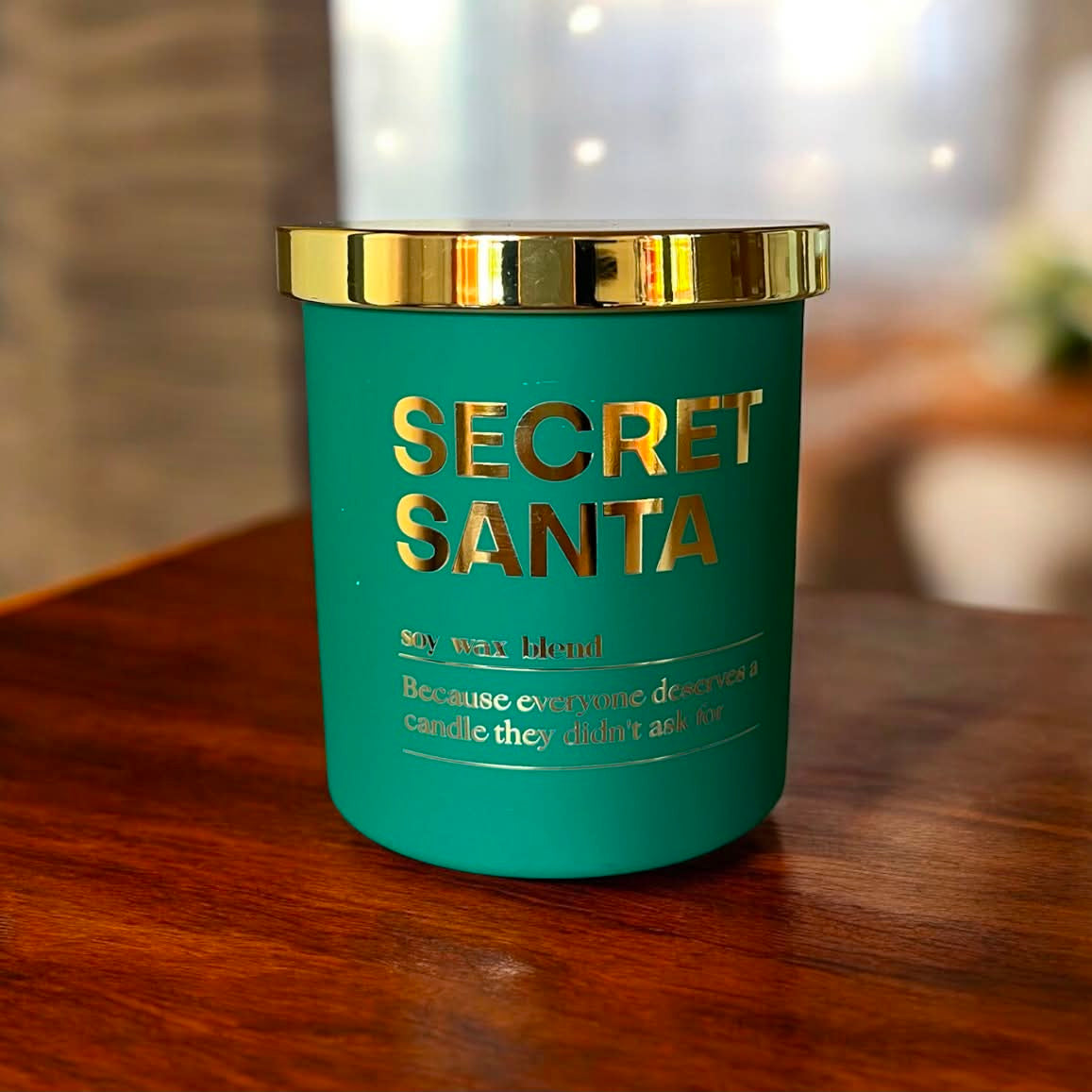 A teal green soy wax candle jar with a shiny gold lid and gold lettering that reads 'SECRET SANTA' on the front.