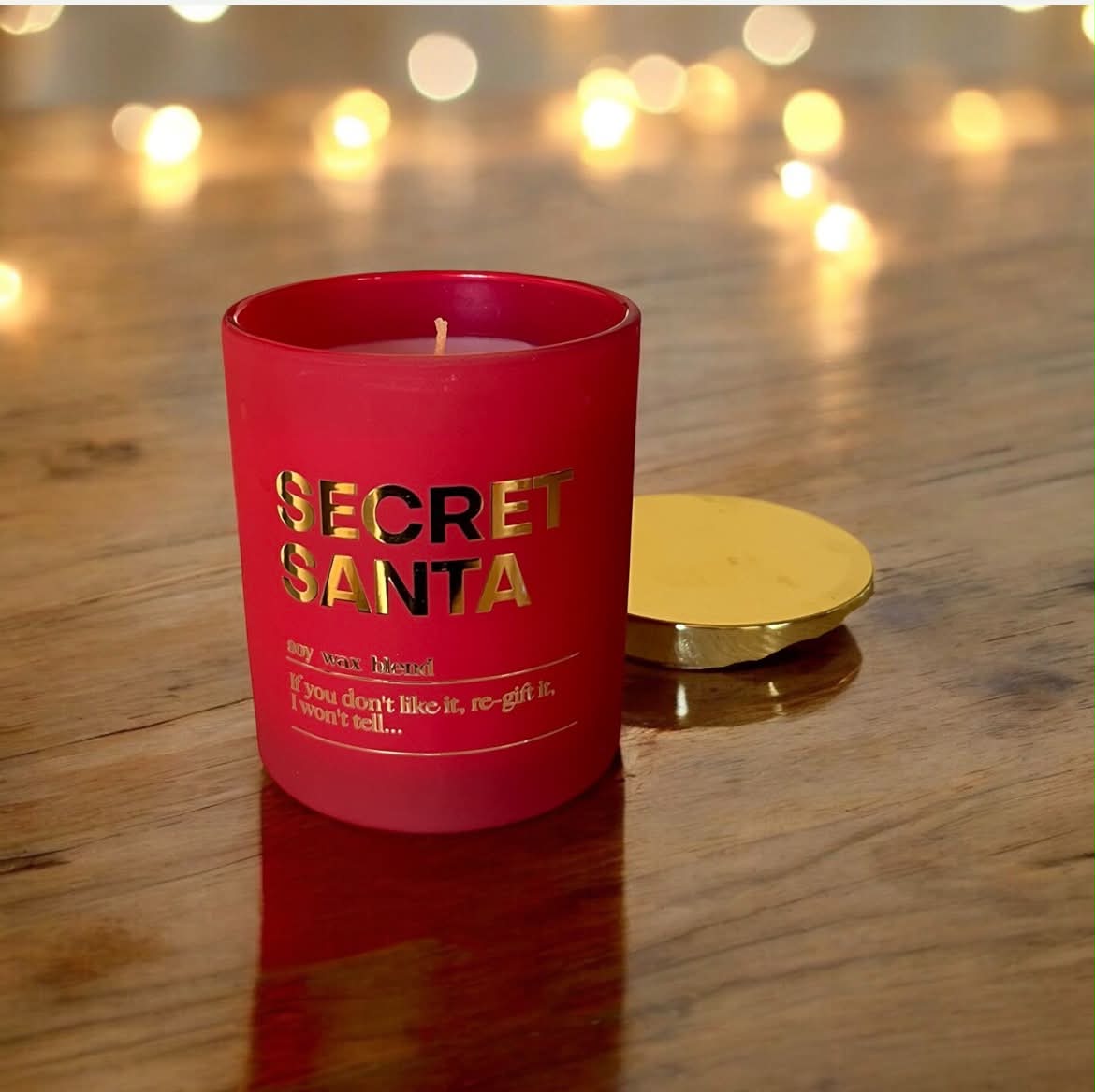 A red glass candle jar with gold foil text reading 'SECRET SANTA' sits on a wooden surface with a gold lid beside it. 