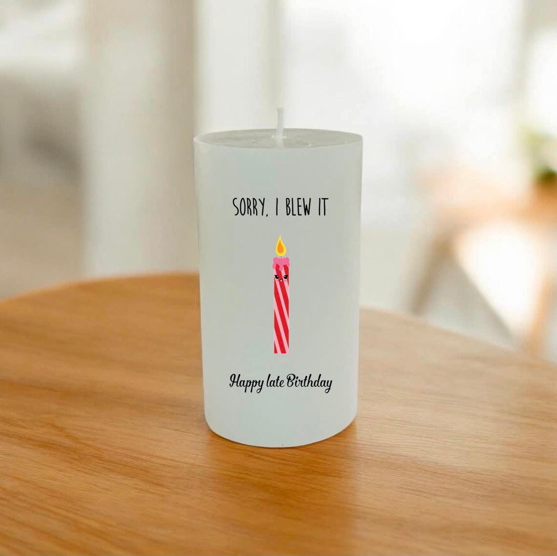 A white candle on a wooden table features a printed graphic of a pink and red striped candle with a yellow flame, accompanied by the text 'SORRY, I BLEW IT' above and 'Happy late Birthday' below.
