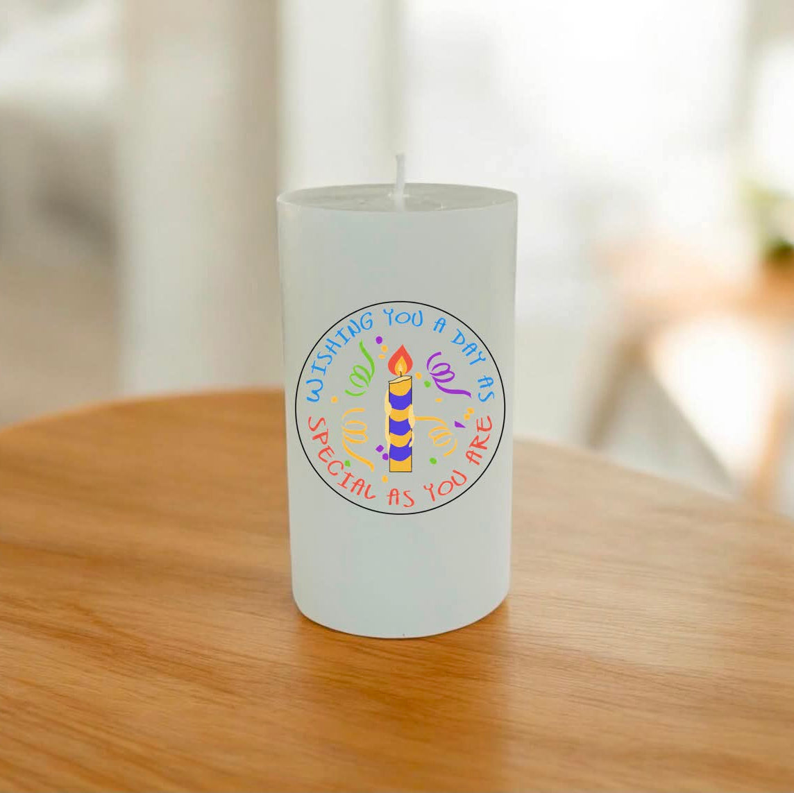 A frosted white cylindrical candle with a colorful circular birthday design on the front, sitting on a wooden table with a softly blurred indoor background.