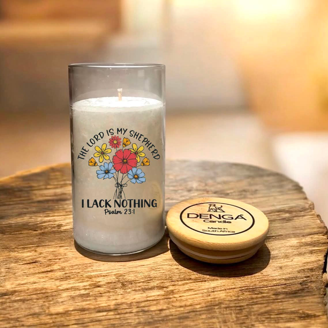 A glass jar candle with a floral graphic and the text 'THE LORD IS MY SHEPHERD I LACK NOTHING Psalm 23:1' sits on a rustic wooden surface next to its round wooden lid.