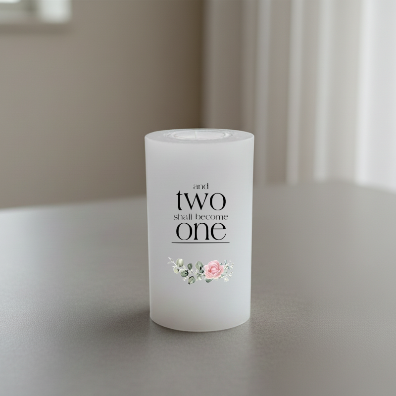 A white cylindrical candle with the printed phrase 'and two shall become one' in black serif text, plus a small pink and white floral illustration near the bottom.